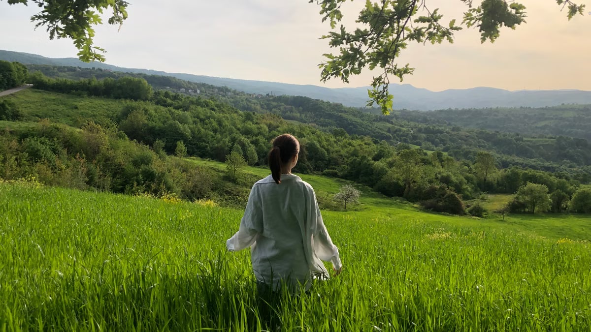 A woman stands in a lush green field, surrounded by rolling hills and forests, enjoying nature's serenity.